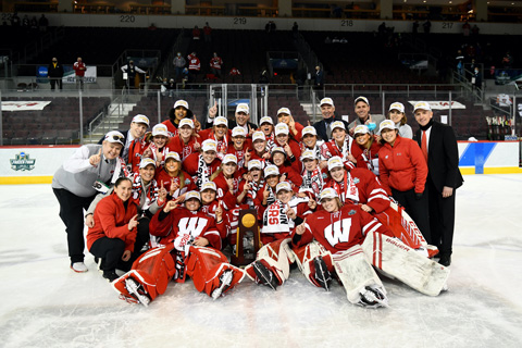 The women's hockey team poses on the ice with coaches and an NCAA championship trophy