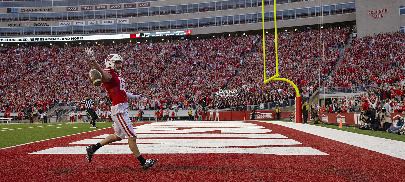 A Wisconsin Badgers football player runs into the end zone flipping the ball in celebration, in front of a packed Camp Randall Stadium crowd.