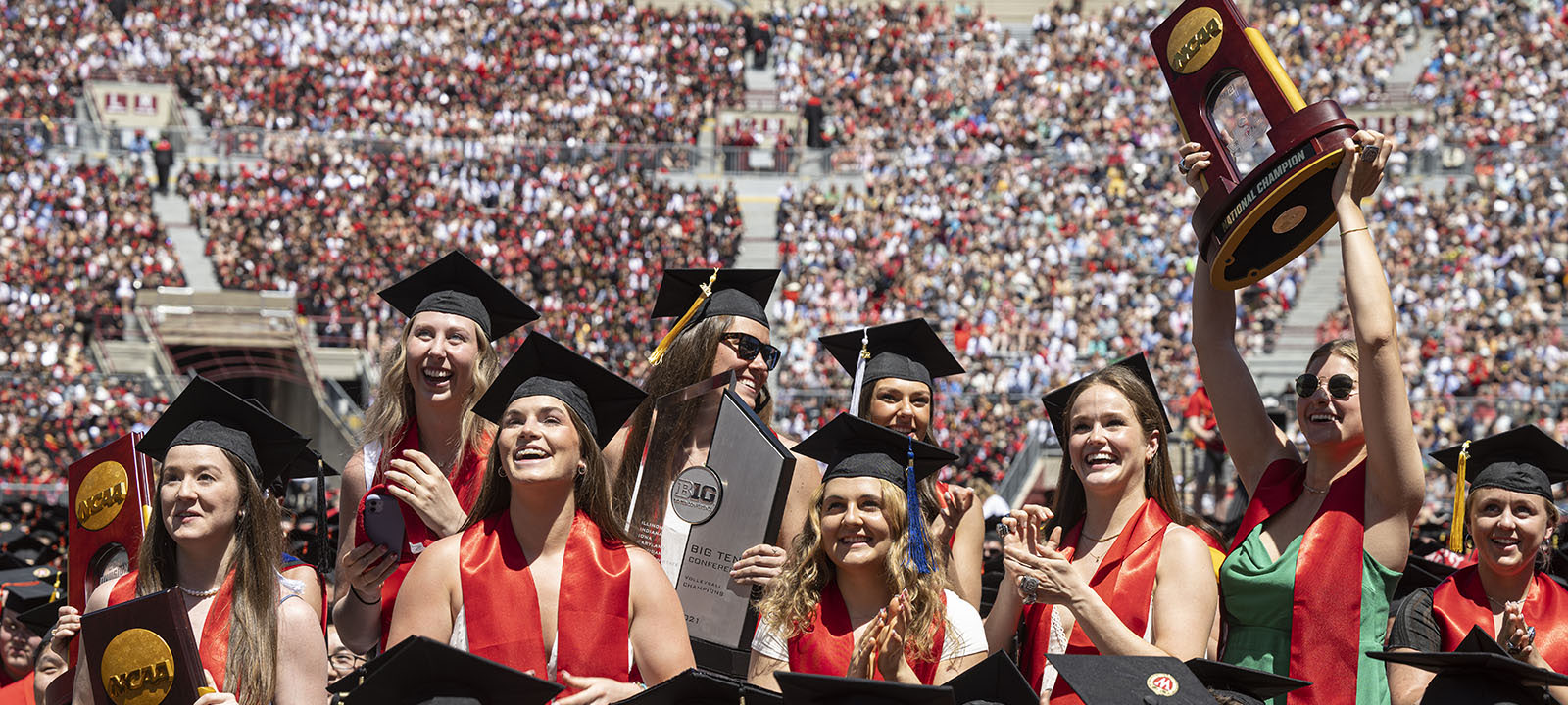 Graduating Badger student-athletes in caps and gowns smile and hold up an NCAA championship trophy at commencement in Camp Randall Stadium.