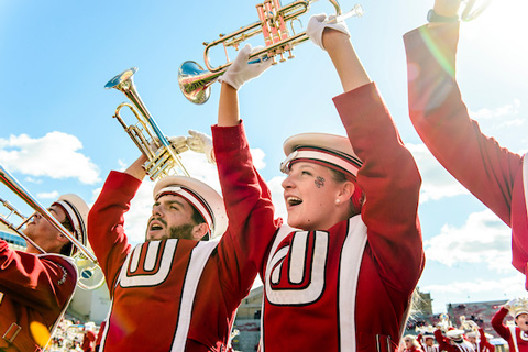 Two members of the UW Marching Band hold their trumpets over their heads during a performance outdoors on a sunny day.