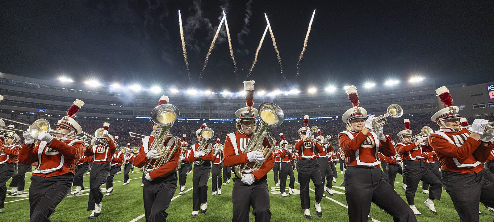 The University of Wisconsin Marching Band performs on the field at Camp Randall Stadium at night with fireworks in a U W shape shooting up behind them.
