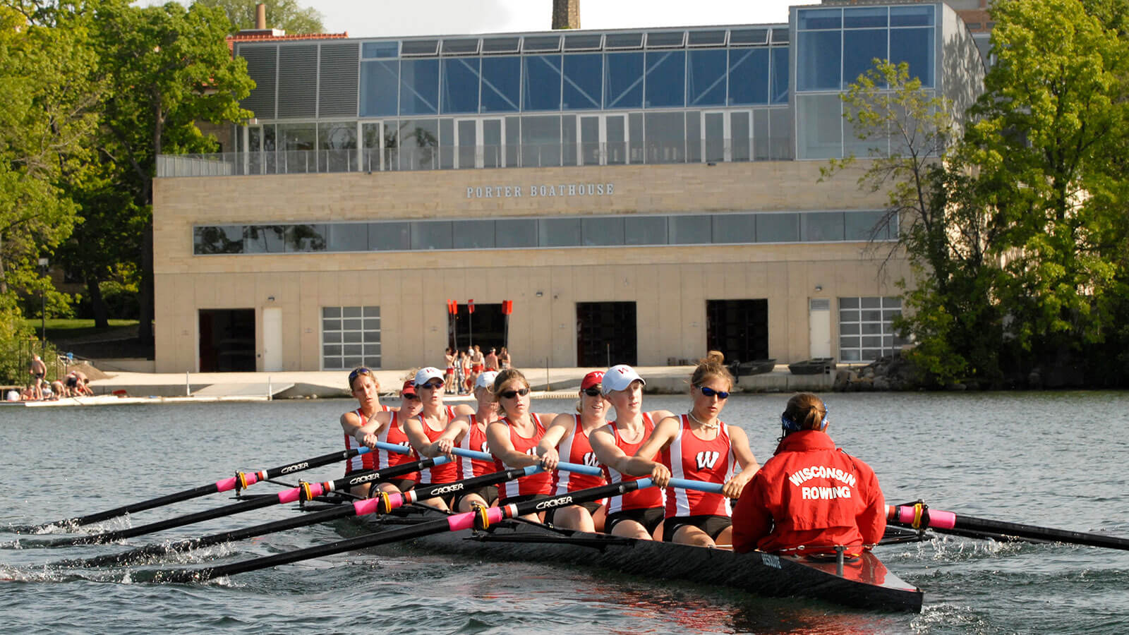 Women's crew team rowing on Lake Mendota with Porter Boathouse in the background.