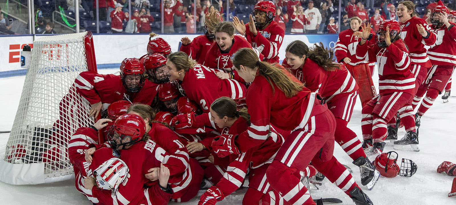 The Wisconsin women's hockey team celebrates a championship win in a jubilant pile in front of the goal on the ice.